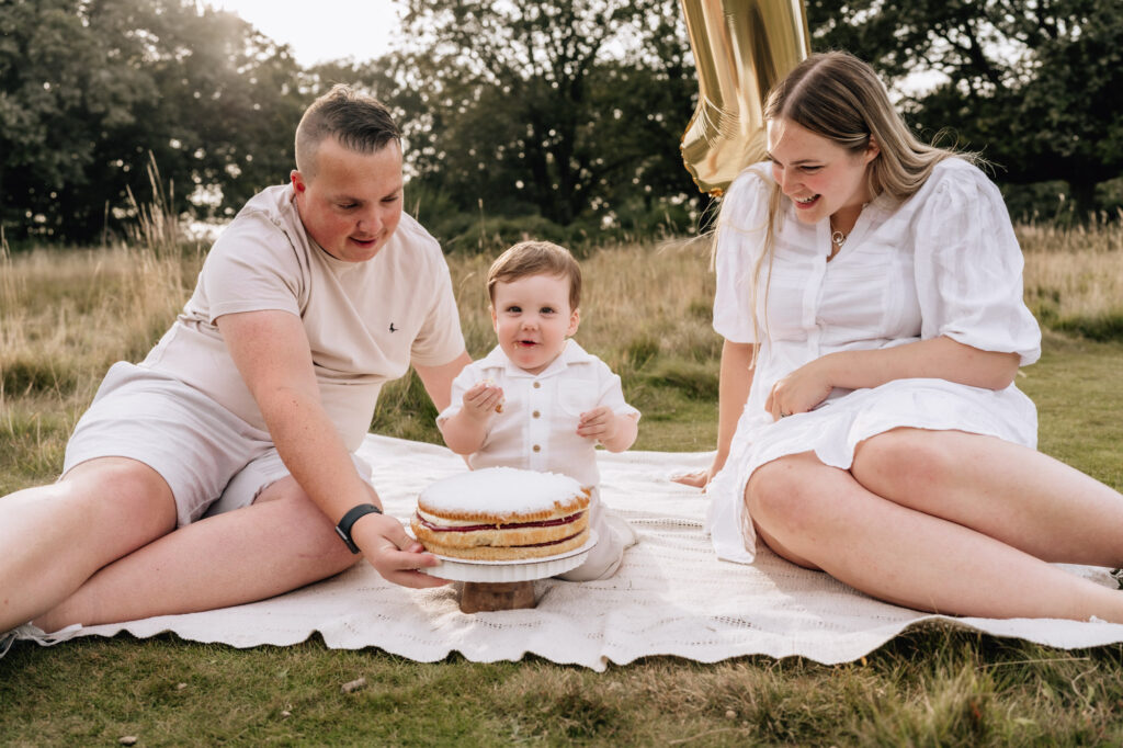 A mum and dad dressed in creams and whites sitting with tier young son on a picnic blanket in a field. They have a tiered cake on a cake stand in front of the boy.